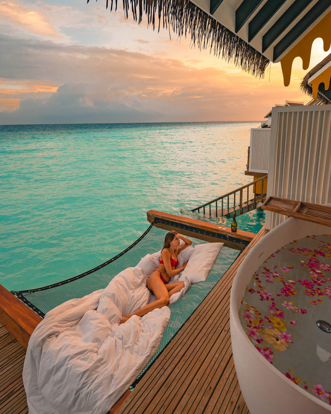girl sitting in a hammock with pillows next to a bath tub with flowers on the terrace of a water villa in The Maldives and watching the sunset