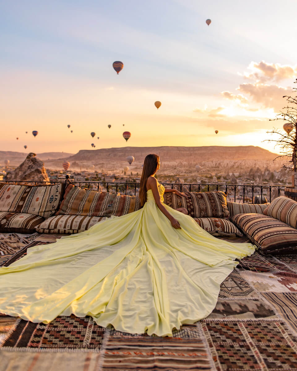 girl with yellow long dress overlooking the sunrise and hot air balloons from the terrace of the Koza Cave Hotel in Cappadocia