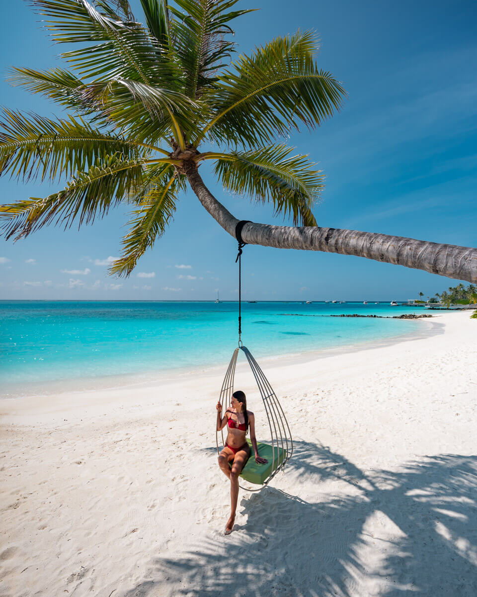 girl sitting on a beach swing at the white and sunny beach, hanging on a palm tree with blue water and sunshine